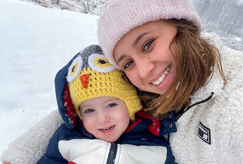 college-aged girl poses with child in jackets and beanies in the snow