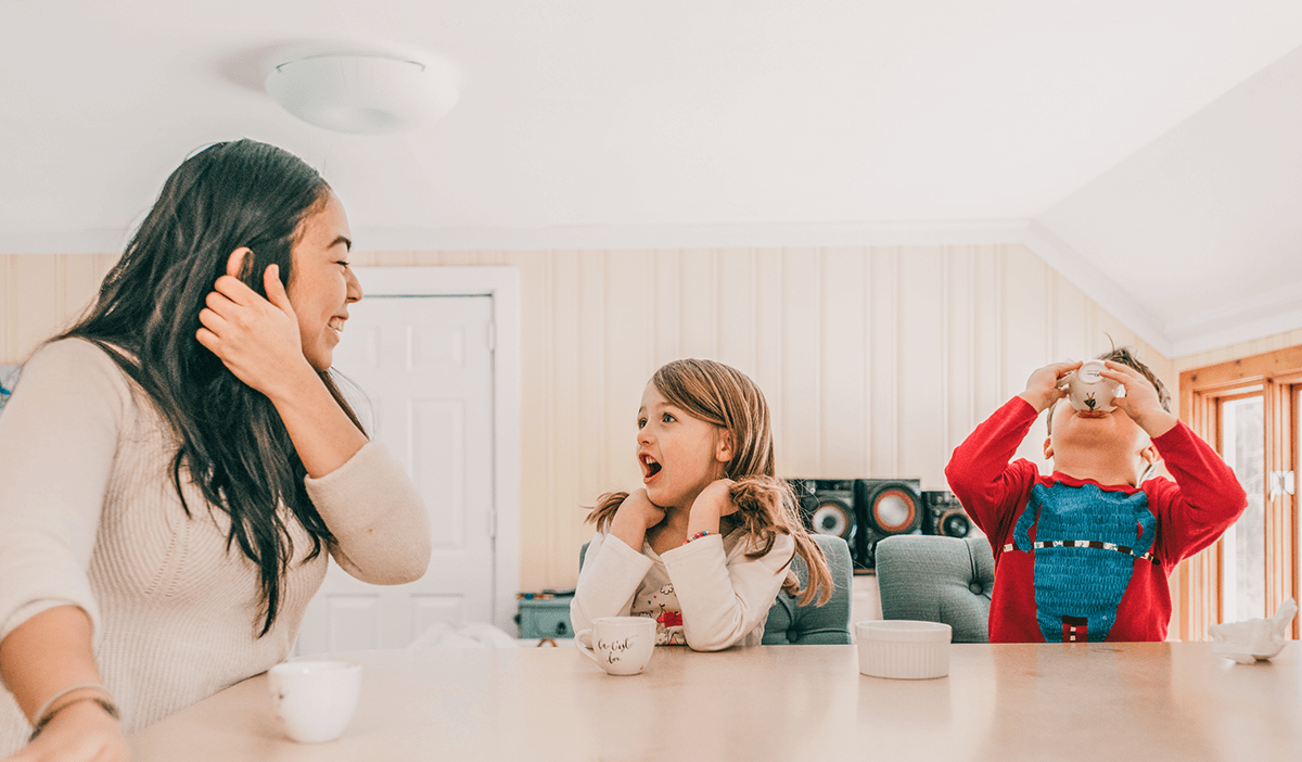 female caregiver is feeding two young children at a table inside of a home